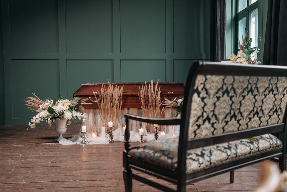 A floral arrangement featuring white roses, pale pink carnations, and greenery in a white vase, placed on a wooden table alongside tall, beige pampas grass. The setting includes ambient lighting with