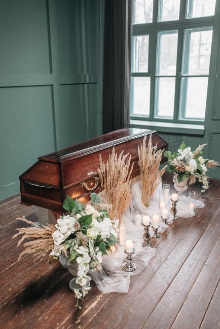 A floral arrangement placed on the floor of a room featuring a vintage wooden coffin with intricate detailing. The display includes two large bouquets of white roses, hydrangeas, and other soft-hued b