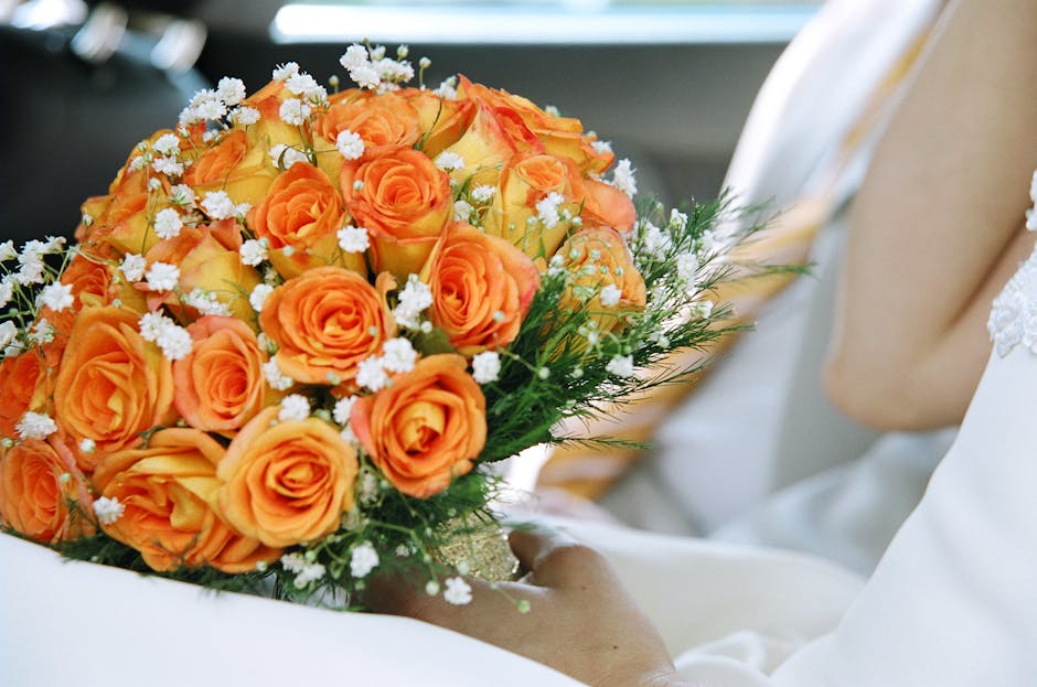A close-up of a floral bouquet featuring vibrant orange roses with tightly curled petals and a velvety texture, complemented by small white filler flowers and delicate greenery. The bouquet is wrapped