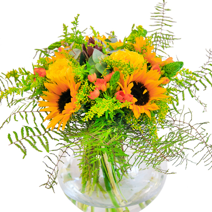 Upon first viewing, this Upminster-inspired bouquet dazzles with a sunlit vibrancy, its radiant hues bringing thoughts of golden afternoons along Cranham Road. Towering, velvety sunflowers command the arrangement with their bold, dark-centered faces, while generous yellow garden roses beam beside them, their multi-layered petals unfolding in classical spirals. Glossy crimson hypericum berries sparkle among the varied foliage, their playful appearance contrasting beautifully with the matte finish of petals and leaves. Lush green elements cascade artfully: feathery fern fronds add a gentle sense of motion, like leaves rustling near Upminster Windmill on a crisp autumn day. Tucked within are airy sprigs of goldenrod, bearing delicate yellow tufts that catch the light, and slender spikes of fresh greenery. Ernst eucalyptus leaves, tinged in blue-green, peek out, bringing a rustic, countryside touch typical of East London's outskirts. The clear, round vase envelops the stems, revealing every vibrant swipe and subtle hue beneath the surface of sparkling water. Up close, one might notice the interplay of silk-soft sunflower petals, tightly wound rose blooms, and crisply bright greens. The scent is subtle but renewing-like a field in late August just after a morning rain. Designed for celebratory moments or as a vibrant accent in any Upminster home, this arrangement channels happiness, optimism, and the gentle artistry of a florist who knows the rhythm of the neighborhood.
